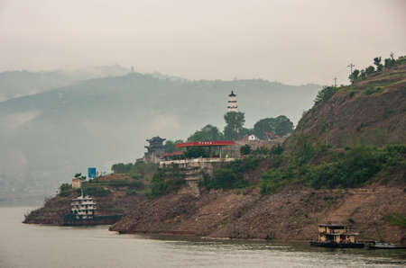 Baidicheng, China - May 7, 2010: Qutang Gorge on Yangtze River. Appearance of historic fortress site on hill with pagoda and entrance gate and towers. White mist over hills in back. Boat on water.のeditorial素材