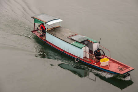 Baidicheng, China - May 7, 2010: Qutang Gorge on Yangtze River. Closeup of Small red motor powered sailing sloop with man and boy against brown water. Red deck and black and green roof.のeditorial素材