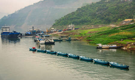 Baidicheng, China - May 7, 2010: Qutang Gorge on Yangtze River. Big blue trasnport boats and row of sloops moored along green agriculture covered shoreline. White mist of mountains as backdrop.のeditorial素材