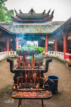 Fengdu, China - May 8, 2010: Ghost City, historic sanctuary. Courtyard with heavy incense vessel and burning sticks. Chinese architecture of halls and roof around under silver sky. Green foliageのeditorial素材