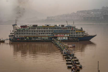 Fengdu, China - May 8, 2010: Morning shot on brown water Yangtze River on passenger boats moored at pontoon bridge of terminal. Skyjline of city buildings under fog and smog.のeditorial素材