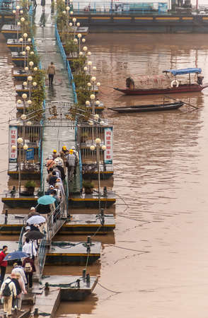 Fengdu, China - May 8, 2010: People walking on narrow pontoon bridge to their boat at Yangtze river terminal. Brown water and a couple of sloops.のeditorial素材