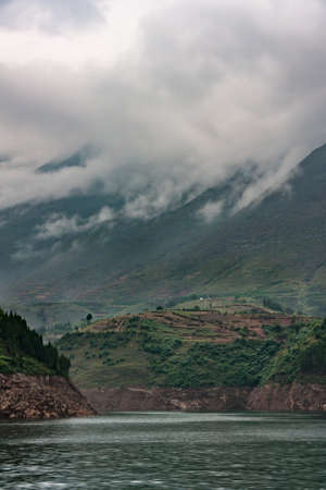 Dragon Gate Gorge on Daning River. Misty cloudscape hide top of mountainous shoreline along emerald green water. Agricultural plots on brown-green hill.の写真素材