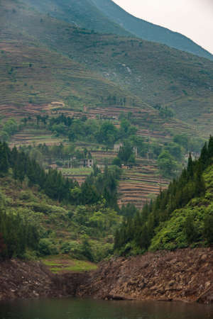 Dragon Gate Gorge on Daning River. Brown dirt Terraced agriculture plots on steep mountain shoreline with plenty of green tree foliage. Some silver sky.の写真素材