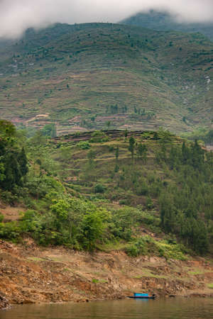 Dragon Gate Gorge on Daning River. Wide portrait of green terraced agriculture plots on steep green mountain with small blue cabin boat on green water at brown dirt shoreline.の写真素材
