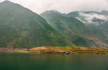 Dragon Gate Gorge on Daning River. Landscape with multiple tall green mountains in misty cloudscape. Some agriculture on shore of emerald green water with boat.の写真素材