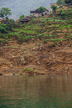 Dragon Gate Gorge on Daning River. Farm house above terraced agriculture plots of green on brown dirt descending in enerald green water. White cable from top.の写真素材