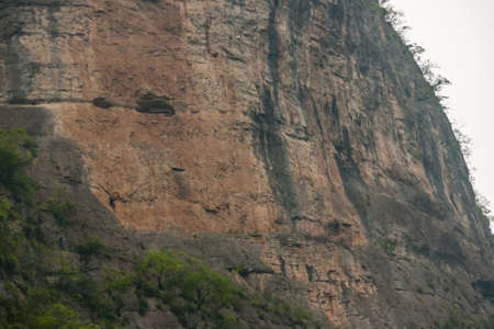 Dawu or Misty Gorge on Daning River. Caves with coffins of ancient tribal people hewn in brown cliff. Some foliage at bottom and piece of silver sky.の写真素材