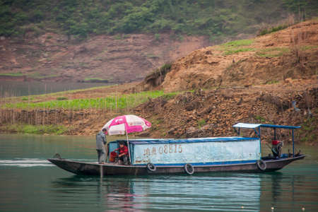Wuchan, China - May 7, 2010: Dragon Gate Gorge on Daning River. closeup of blue cabin boat with 2 people and pink umbrella on emerald green water in front of brown dirt agriculture plot.のeditorial素材