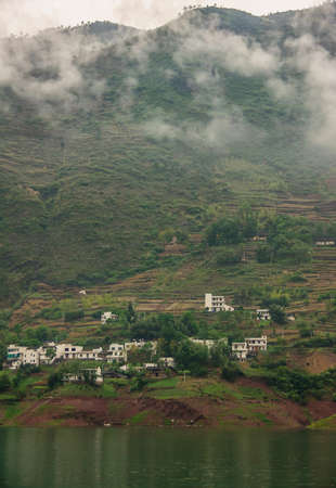 Wuchan, China - May 7, 2010: Dicui or Emerald Gorge on Daning River. White houses of village set on greem mountain slope with terraced agriculture plots above dark green water and mist above.の写真素材