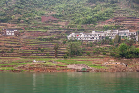 Wuchan, China - May 7, 2010: Dicui or Emerald Gorge on Daning River. White houses of village set on brown terraced agriculture plots above green water. Green vegetation.の写真素材