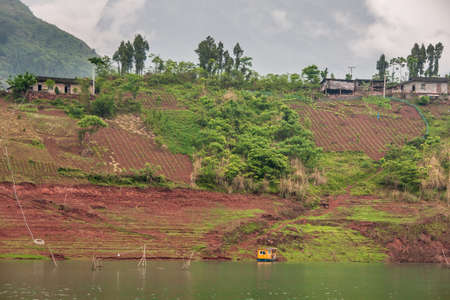 Wuchan, China - May 7, 2010: Dicui or Emerald Gorge on Daning River. Brown farmland with green vegetation on slope descending to green river with fish pen, and cabin boat under cloudscape.の写真素材