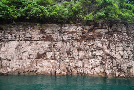 Wuchan, China - May 7, 2010: Dragon Gate Gorge on Daning River. Closeup of beige-brown rocky shoreline covered with brown dirt and green foliage on top, descending in emerald green water.の写真素材