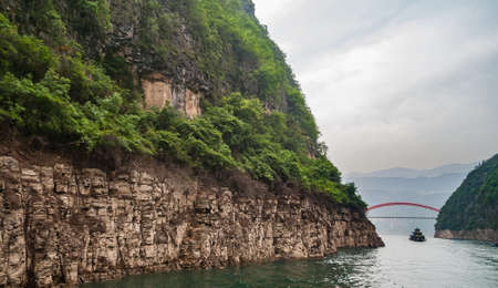 Wuchan, China - May 7, 2010: Dragon Gate Gorge on Daning River. Red bow with flat white concrete road surface S103 bridge over green water at end of canyon under silver sky. Steep rocky shorelines with green foliage.の写真素材