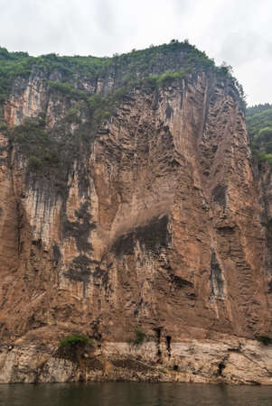 Wuchan, China - May 7, 2010: Dragon Gate Gorge on Daning River. Tall brown and black cliff descending into dark green water. Green foliage on top and flank. Silver sky.の写真素材