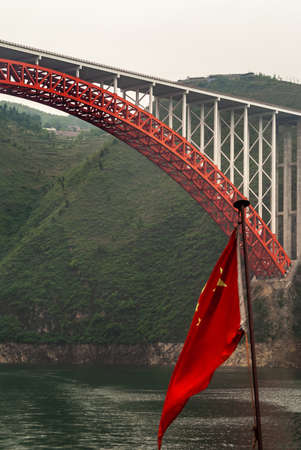 Wuchan, China - May 7, 2010: Dragon Gate Gorge on Daning River. Red bow with flat white concrete road surface bridge over dark green water and Chinese flag. Mountainous shorelines with green foliage.の写真素材