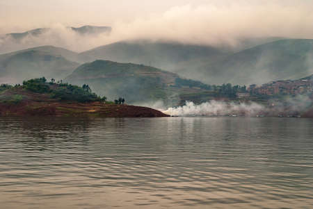 Wuchan, China - May 7, 2010: Dawu or Misty Gorge on Daning River. Wide landscape over water showing agriculture land on hills and green mountains under cloudscape. Fire smoke at edge.の写真素材