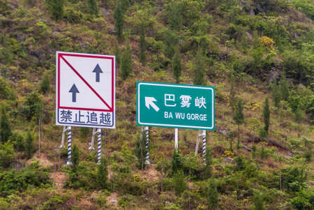 Wuchan, China - May 7, 2010: Dawu or Misty Gorge on Daning River. Boat traffic signs on steep green foliage hill. Ba Wu or Dawu white on green. Blue and red directional arrows on white.の写真素材
