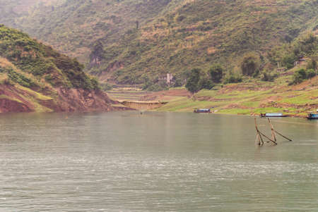 Wuchan, China - May 7, 2010: Dicui Gorge on Daning River. Landscape with inham squeezed between agriculture land and mountains, with sticks of fish pen in front in gray-green water.の写真素材