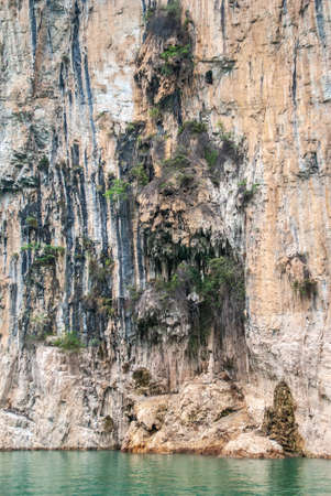 Wuchan, China - May 7, 2010: Dawu or Misty Gorge on Daning River. Packs of stalactites hang form brown cliffs in front of caves above emerald geen water.の写真素材