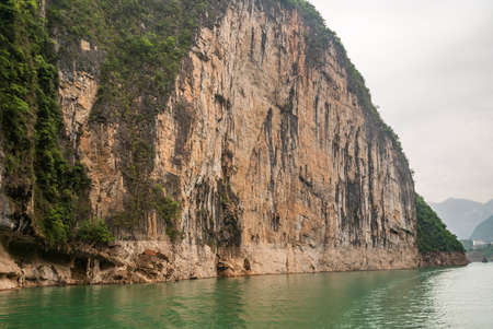 Wuchan, China - May 7, 2010: Dawu or Misty Gorge on Daning River. Huge brown Cliff wall descends into emerald green water, with hanging coffins. Green foliage and silver sky.の写真素材