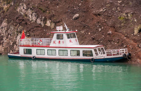Wuchan, China - May 7, 2010: Dicui or Emerald Gorge on Daning River. Small white passenger boat moored along brown dirt mountain slope sitting on green emerald water. Chinese flag.のeditorial素材