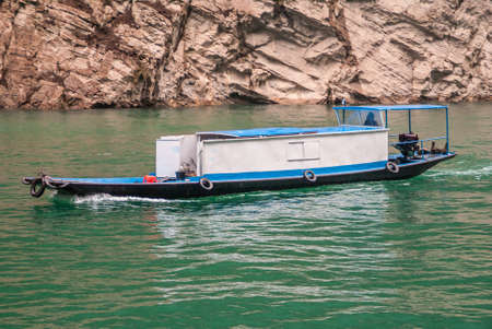 Wuchan, China - May 7, 2010: Dawu or Misty Gorge on Daning River. Closeup of small White-blue cabin boat travels on emerald green water with brown rocky cliff as backdrop.のeditorial素材