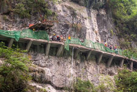 Wuchan, China - May 7, 2010: Dawu or Misty Gorge on Daning River. Plank pathway is turned into concrete by work crew with green portective fence built hanging on brown-black bare cliff..のeditorial素材