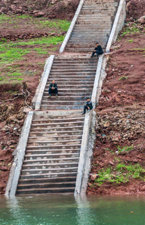 Wuchan, China - May 7, 2010: Dicui Gorge on Daning River. 3 ferry passengers wait on long steep stairway on mountain slope above emerald green water. Brown dirt around.のeditorial素材