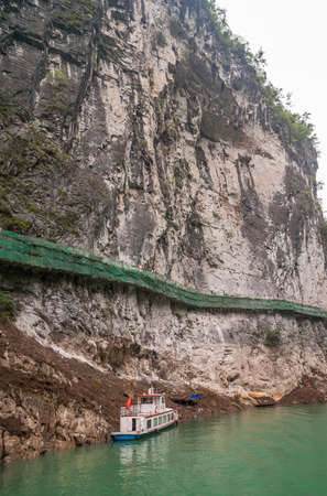 Wuchan, China - May 7, 2010: Dawu or Misty Gorge on Daning River. Boat and plank pathway with green portective fence built hanging on brown-black bare cliff above emerald green water.の写真素材