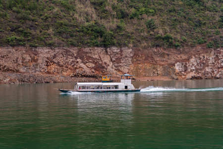 Wuchan, China - May 7, 2010: Dicui Gorge on Daning River. White ferry boat with passengers speeding on emerald green water in front of brown rocky green folliage covered shoreline with slipway.の写真素材