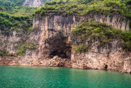 Wuchan, China - May 7, 2010: Dicui Gorge on Daning River. Deep cave in brown-black rocky cliff with green foliage on top. Emerald green water up front,の写真素材