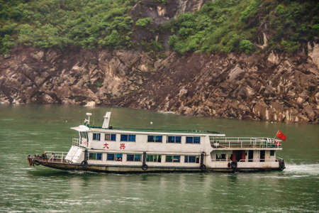 Baidicheng, China - May 7, 2010: Qutang Gorge on Yangtze River. Old rusty white passenger ferry on green water with brown rocky shoreline and some green vegetation on top. Red flag.のeditorial素材