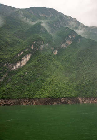 Baidicheng, China - May 7, 2010: Qutang Gorge on Yangtze River. Gray cloudscape descends along slopes of green covered mountain in back of green water. Some beige brown cliffs.の写真素材