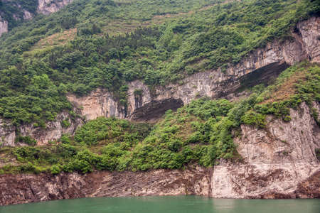 Zigui, China - May 6, 2010: Xiling gorge on Yangtze River. Large caves in brown rock cliffs with green foliage on top above green water.の写真素材