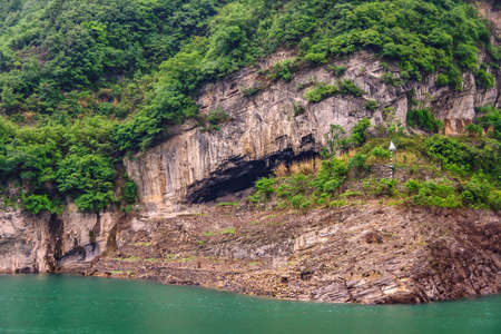 Zigui, China - May 6, 2010: Xiling gorge on Yangtze River. Large cave in brown rock cliff with green foliage on top above green water.の写真素材