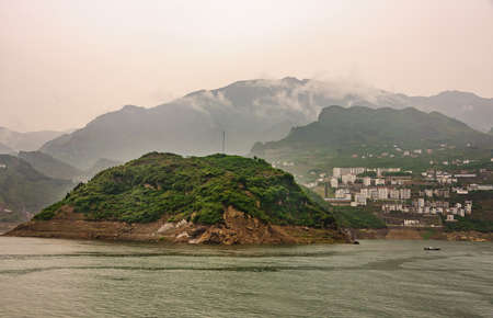 Xiangxicun, China - May 6, 2010: Xiling gorge on Yangtze River. Wide landscape with green hill and Xiangxicun town houseing in back. Green mountains and gray descending cloudscape as backdrop.の写真素材