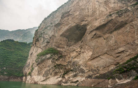 Xiangxicun, China - May 6, 2010: Xiling gorge on Yangtze River. Large cave hole in brown rock cliff descending in green water. Green covered mountains as backdrop. Patch of silver sky.の写真素材