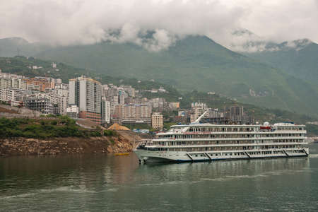 Xinling, China - May 6, 2010: Xiling gorge on Yangtze River. President cruise boat on green water in front of highrise section of city with descending cloudscape along flanks of green mountain range.のeditorial素材
