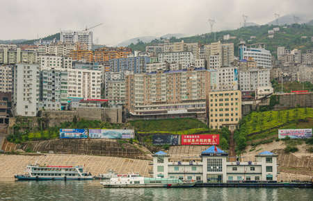 Xinling, China - May 6, 2010: Xiling gorge on Yangtze River. Police building on shoreline with boats. Wall of tall buildings as backdrop. Advertesement billboards add colors. Gray cloudscape.のeditorial素材