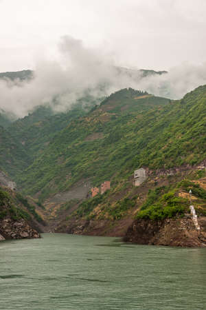 Shengli Street, China - May 6, 2010: Xiling gorge on Yangtze River. Houses built on edge of slope of  green tree covered mountain slope above green water. Descending cloudscape.の写真素材