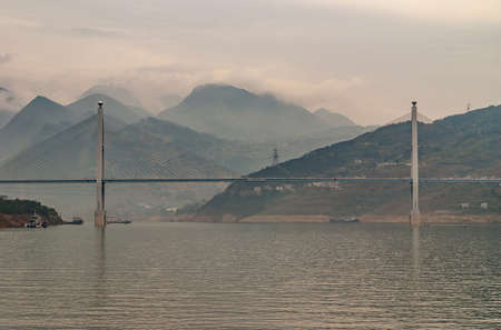 Xinling, China - May 6, 2010: Xiling gorge on Yangtze River. Badong Suspension Bridge with its 2 pylons over wide green water. Mountains and cloudscape in back. Some boat traffic.の写真素材