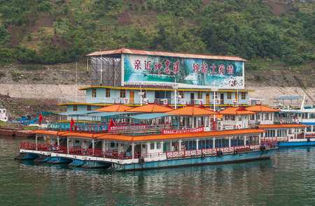 Xinling, China - May 6, 2010: Xiling gorge on Yangtze River. Local short river cruise ofice building termina and boatsl on green water. Green mountain slope as backdrop. Colorful billboard.のeditorial素材