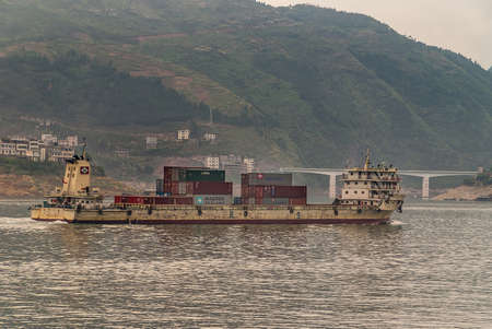 Xinling, China - May 6, 2010: Xiling gorge on Yangtze River. Beige rusted shipping container boat with tall green mountains in back, and road bridge.のeditorial素材
