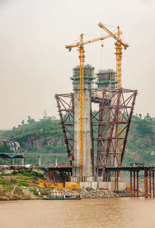 Fengdu, Chongqing, China - May 8, 2010: Yangtze River. Closeup of concrete suspension tower for bridge under construction with 2 tall yellow cranes over brown water, silver sky and green hill.の写真素材