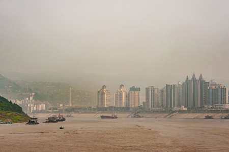 Fengdu, Chongqing, China - May 8, 2010: Yangtze River. Cityscape with tall buildings on shore of brown water upon serveral boats. Green mountains in fog and smog.の写真素材