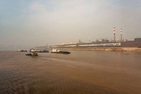 Chongqing, China - May 8, 2010: Yangtze River. Giant power plant and factories on shoreline behind wide brown water with several coal transporting boats under light blue sky.の写真素材