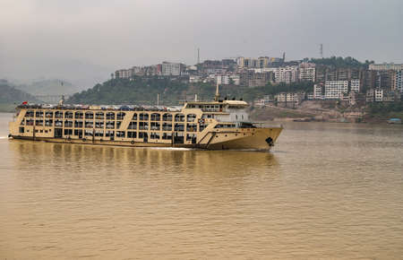 Myaoyinan, Chongqing, China - May 8, 2010: Yangtze River. Yellow new car transport boat on brown water with cityscape in back under dark gray sky.のeditorial素材