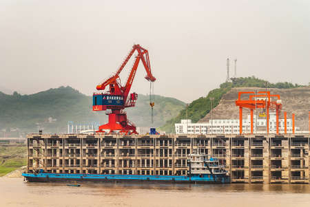 Huangqikou, Chongqing, China - May 8, 2010: Yangtze River. Red and blue crane unloads blue barge at multi-level quay of port. Green hill under smog sky in back, Brown water up front.の写真素材