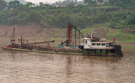 Huangqikou, Chongqing, China - May 8, 2010: Yangtze River. Ongoing dredging operation of 2 boats. Small Pumping vessel behind barge filled with heaps of wet dirt. Green shoreline with housing on top.の写真素材